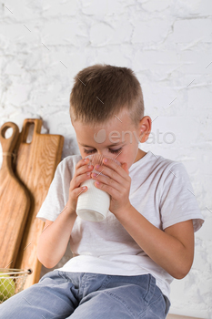 A blond boy in a white T-shirt drinks milk from a tumbler in the kitchen.