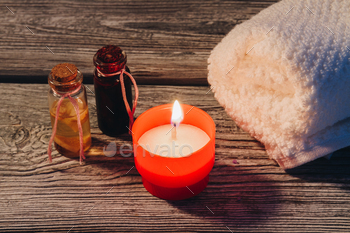 Bottle of oil with candle and white rolled towel on wood.