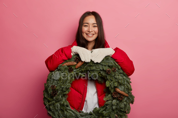 Brunette cheerful younger Asian woman with lengthy hair, natural beauty, leans at inexperienced festive wreath, h