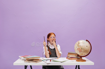 Little ginger girl with pigtails in glasses and grey sundress yawning and sitting at desk with books