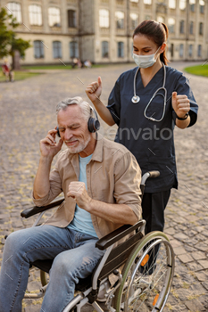 Younger nurse in protective conceal taking care of senior recuperating patient in wheelchair carrying