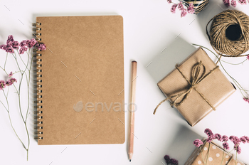 Gift box wrapped in kraft paper, notebook and red plant life on white background. Flat lay styling.