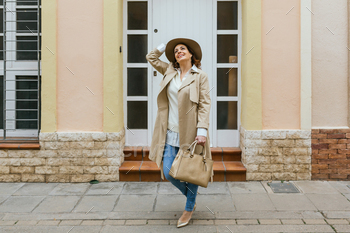 An adult lady in front of her front door posing with her hat and purse whereas taking a look up