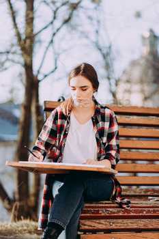 Younger lady artist draws a image while sitting on a bench.