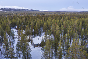 Aerial of picket cabin within the remote forest, with snow coated woods and trees landscape and mountai