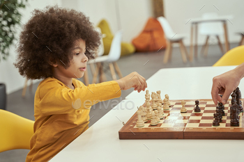 Gorgeous minute suave boy going to secure a transfer while playing chess with grownup, sitting on the table