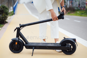 Unrecognizable girl in a white swimsuit folding an electrical scooter within the boulevard