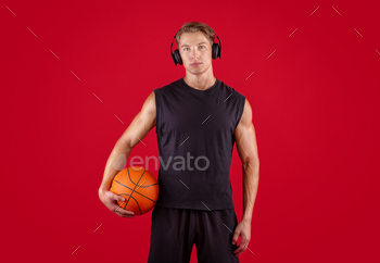 Horny millennial basketball participant being attentive to music, wearing headphones over crimson studio