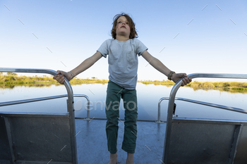 A boy on a motorboat travelling along a waterway, standing holding on to railings.