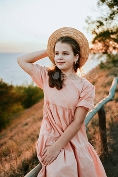 Teen lady wearing pastel costume and straw hat sitting on picket fence exterior in golden sundown light