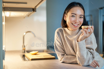 Portrait of smiling younger tickled lady staying at dwelling, standing in kitchen and eating toast