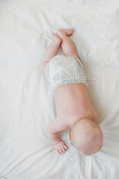 The little one lies on her stomach on a white sheet on the bed stare from above. Newborn little one in diaper