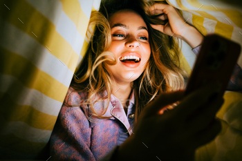 Young cheerful caucasian lady the utilize of smartphone, lies under a blanket on a bed at home