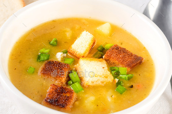 Split pea soup with croutons and green onion in a bowl on white desk.