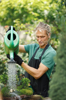 Younger blonde man watering a plant in a pot at yard backyard