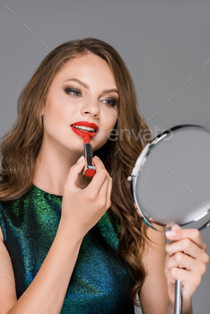 portrait of magnificent younger lady wanting at deem while applying crimson lipstick isolated on gray