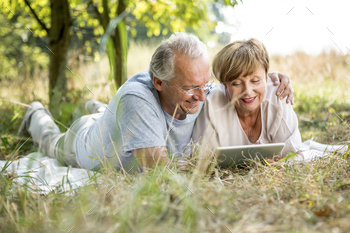 Chuffed senior couple with tablet lying in meadow