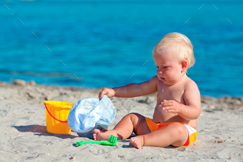 Toddler sitting on the seashore and fiddling with plastic toys within the sand.