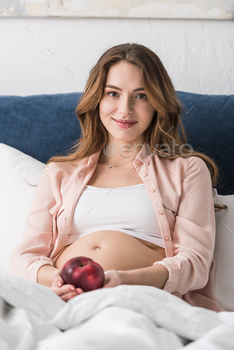 Chuffed pregnant lady lying in mattress with apple and taking a gaze at digicam