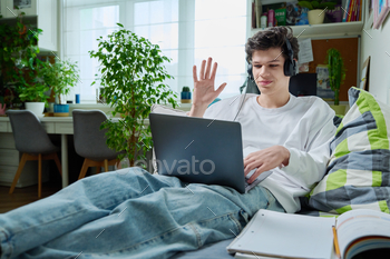 Younger guy in headphones the utilize of computer for video conversation, lying at dwelling