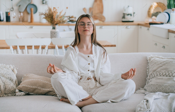 Teen lady in informal sitting in sofa in lotus pose practicing meditation eyes closed at residence