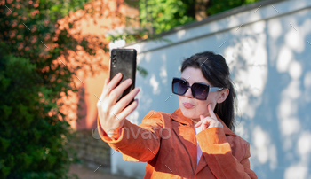 a young brunette woman in an orange jacket takes a selfie in opposition to a brick wall