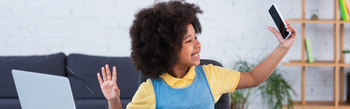 Smiling african american baby waving hand at pc pc and keeping smartphone at dwelling, banner