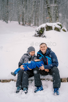 Diminutive lady lies on the knees of smiling mom and pa and catches snowflakes in her mouth in the