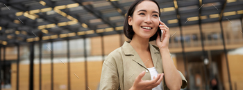 Young asian lady smiles, talks on cell phone and walks in city centre, poses outdoors