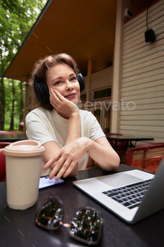 Lovable historic woman with pc and wireless headphones sitting at the table in outdoor cafe