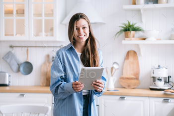 Wonderful brunette lady preserving tablet appears to be like at digicam standing at kitchen relaxing at home. Young woma