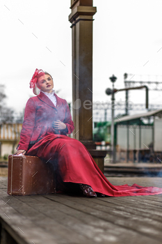 A lady in a burgundy swimsuit and a hat with a veil is sitting on a suitcase discontinuance to an historical locomotive