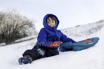 Jubilant child in heat cool local weather attire sitting in snow with a sled.