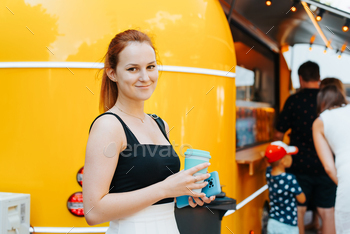 Smiling red-haired younger lady retaining cup of drink whereas standing shut to yellow meals truck in park