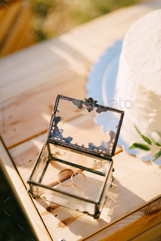 Pair of wedding ceremony rings lies in a pitcher area subsequent to a marriage ceremony cake on a wood area