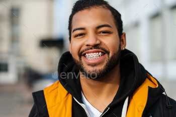 African American scholar with dental braces, carrying jacket, posing on avenue and taking a examine digital camera