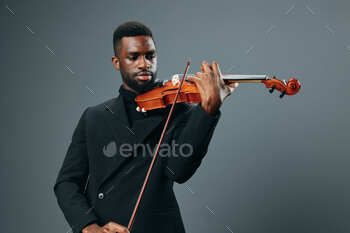 African American man in a shadowy go well with having fun with violin on gray background, final classical musician