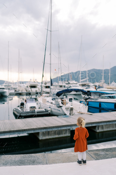 Small woman stands on the pier of a luxurious marina and appears on the rows of moored yachts. Encourage behold