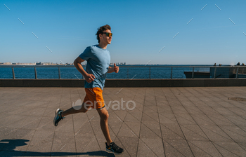 Man jogging alongside a waterfront creep, carrying a blue t-shirt, orange shorts, and reflective sung