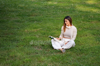 Lady learning a ebook whereas sitting on the grass in a park with headphones.