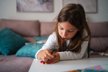 A younger girl focuses on portray her nails whereas preserving a chocolate deal with in a comfortable dwelling