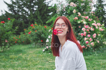 Teenage woman with crimson hair and glasses smiling with a rose in her mouth in a backyard