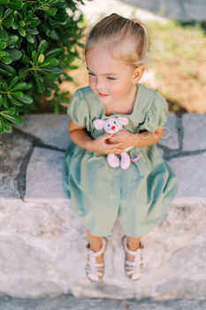 Minute girl sits on a stone backyard fence hugging a purple clean toy mouse