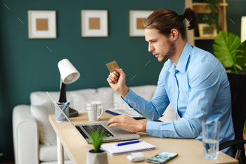 Younger man sitting at a desk with a laptop computer in entrance of him and holds a bank card in his hand