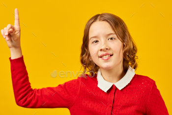 Teenage Schoolgirl in Crimson Cardigan Pointing Upwards In opposition to a Yellow Background