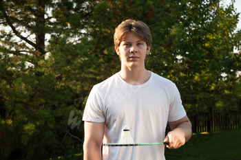 Youthful man retaining white badminton shuttlecock and badminton racket. Summer time sports activities sport. Exterior.