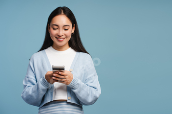 Smiling Asian lady conserving cellular cell phone, utilizing cellular app, standing isolated on blue background