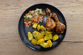 Two roasted rooster drumsticks on a plate with potatoes and boiled greens.